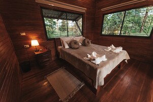 Un dormitorio de cabaña de madera con cama, mesita de noche, lámpara y ventanas amplias con vistas a un bosque.