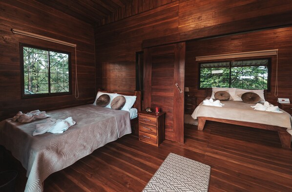 A wooden cabin bedroom with two beds, a nightstand, and a window with a view of trees.