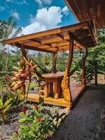 Un espacio de descanso al aire libre con mesas y bancos de madera, rodeado de exuberante vegetación y un cielo azul despejado.