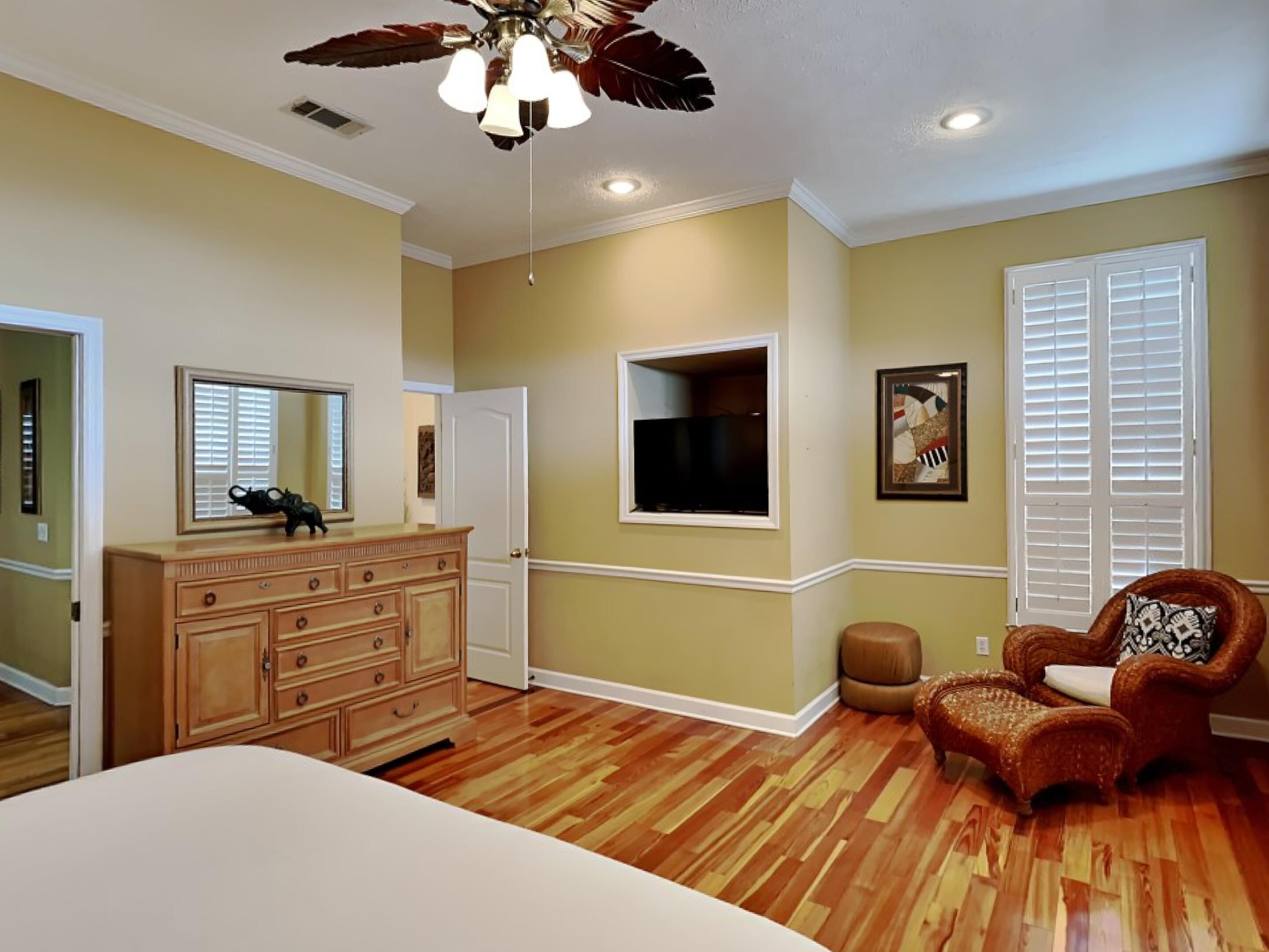 A bedroom with a wooden dresser, a chair, a bed, a ceiling fan, and a window with blinds.