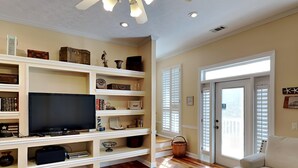 A living room with a flat-screen TV, wooden flooring, white built-in shelves, a white sofa, and a sliding glass door. - Camellia House by Southern Vacation Rentals (Santa Rosa Beach)