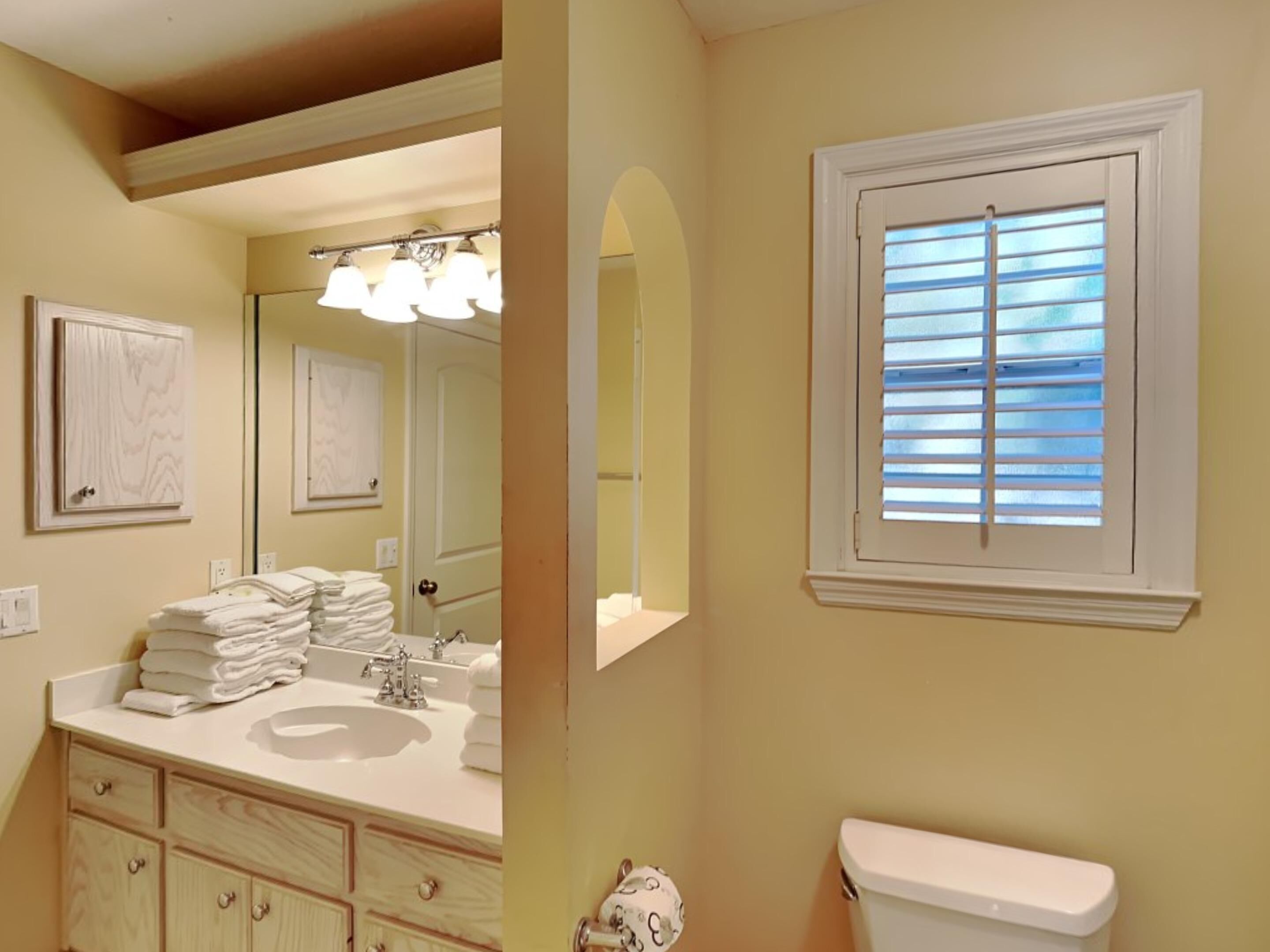 A bathroom with a sink, mirror, and toilet. There is a window with blinds and a towel rack with folded towels.