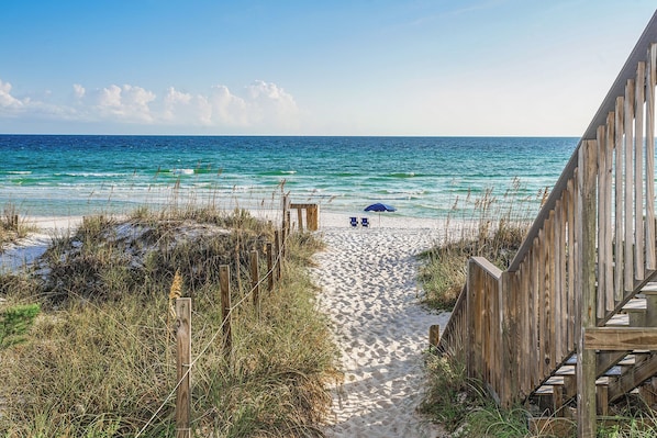 A sandy beach path leading to the ocean, with two chairs and a blue umbrella under a wooden fence.