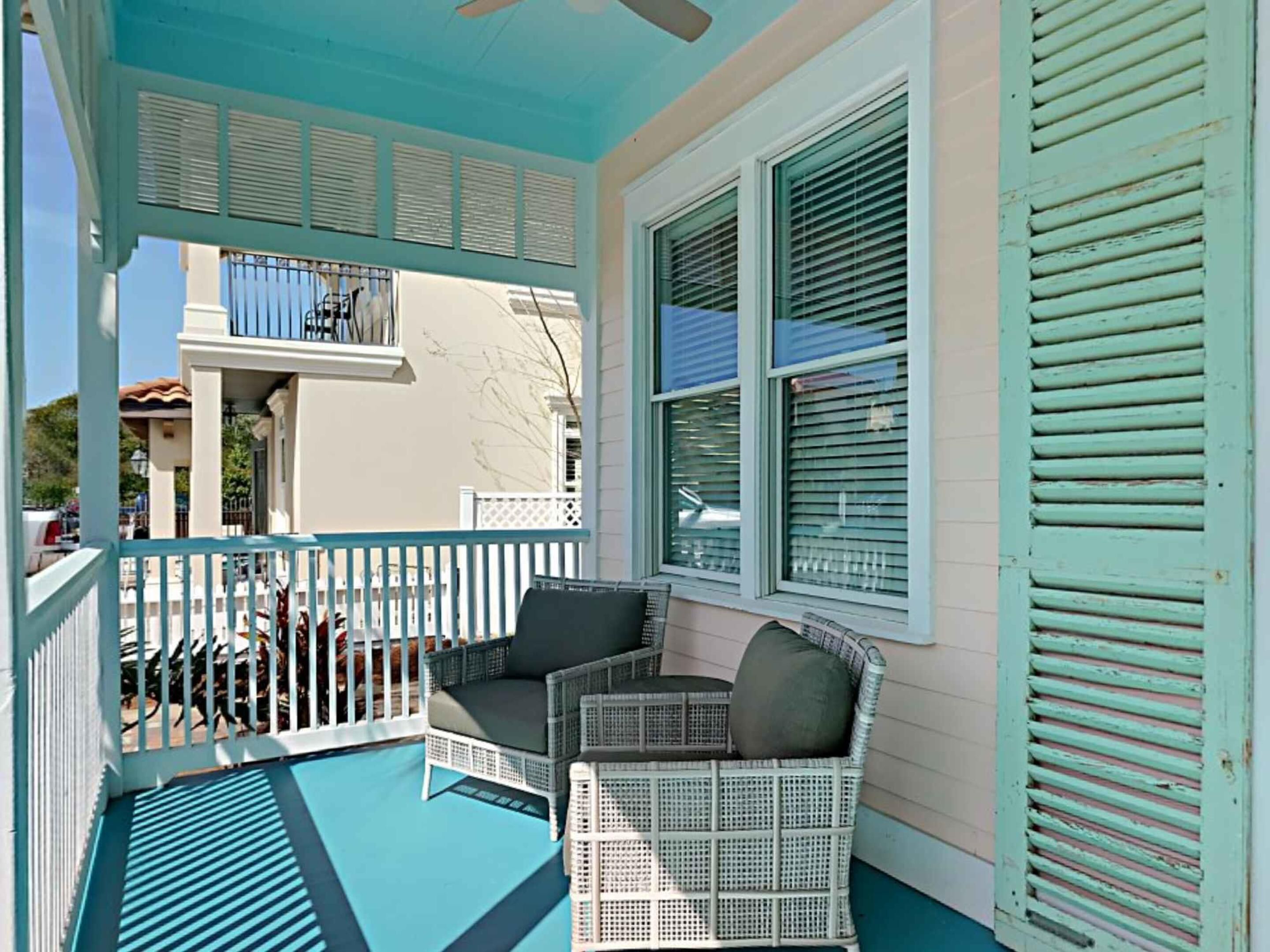 A covered porch with a white wicker chair and a table, a balcony with a railing, and a building with shutters.