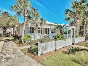 A coastal-style house with a covered porch, palm trees, and a white picket fence.