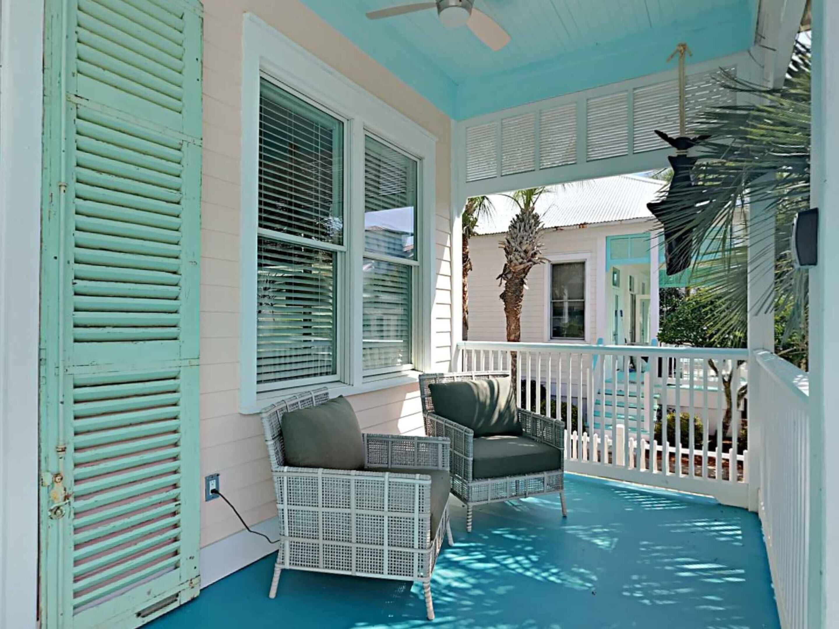 Eine überdachte Veranda mit weißer Reling, ein Korbstuhl mit Kissen und Blick auf ein Haus mit blauer Tür.