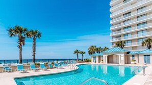 A swimming pool with lounge chairs and palm trees, surrounded by a building with balconies. - Bluewater #403 by Southern Vacation Rentals (Orange Beach)