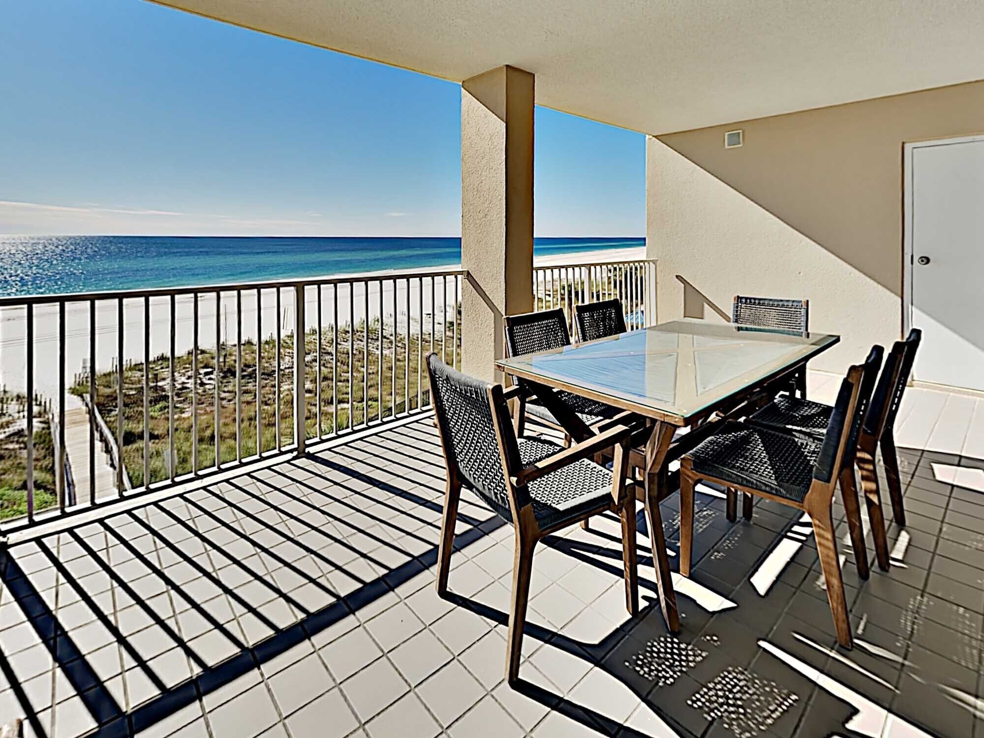 A balcony with a glass table and chairs, overlooking a beach and ocean.