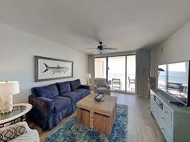 Una sala de estar moderna con un sofá azul, una mesa de centro y un televisor. Hay un balcón con vistas al océano.