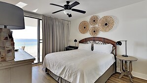 A bedroom with a bed, a ceiling fan, a nightstand, a lamp, and a view of the ocean. - Bluewater #403 by Southern Vacation Rentals (Orange Beach)