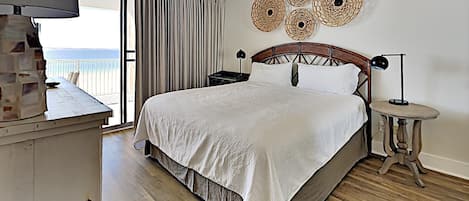 A bedroom with a bed, a ceiling fan, a nightstand, a lamp, and a view of the ocean.