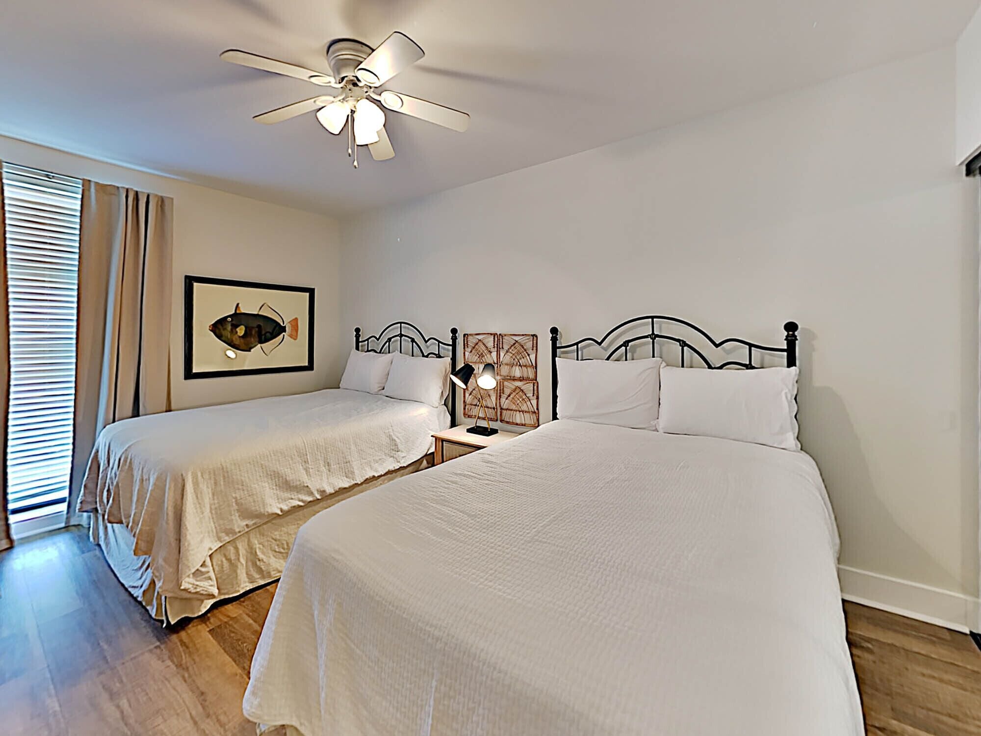 A hotel room with two single beds, a ceiling fan, a framed picture on the wall, and a bedside table with a lamp.