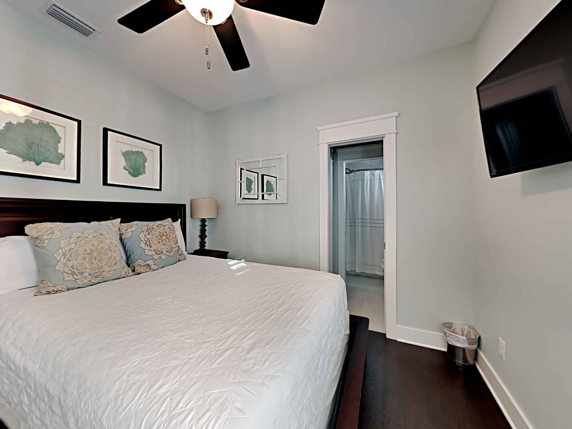 A bedroom with a large bed, a ceiling fan, framed artwork on the wall, a TV mounted on the wall, and a door leading to another room.