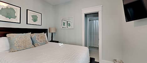A bedroom with a large bed, a ceiling fan, framed artwork on the wall, a TV mounted on the wall, and a door leading to another room.