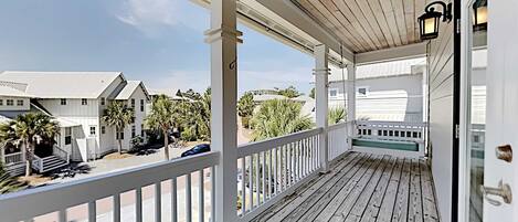 A covered porch with white railings and a wooden deck, overlooking a residential area with houses and palm trees.