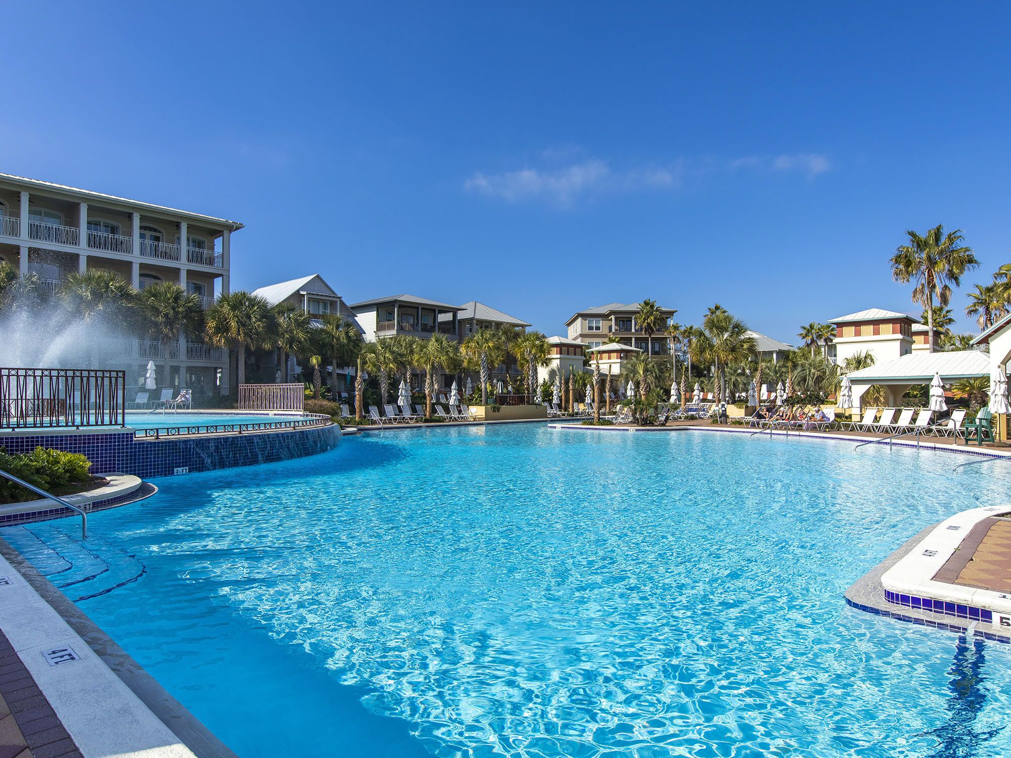 A large outdoor swimming pool with lounge chairs and palm trees, surrounded by hotel buildings.