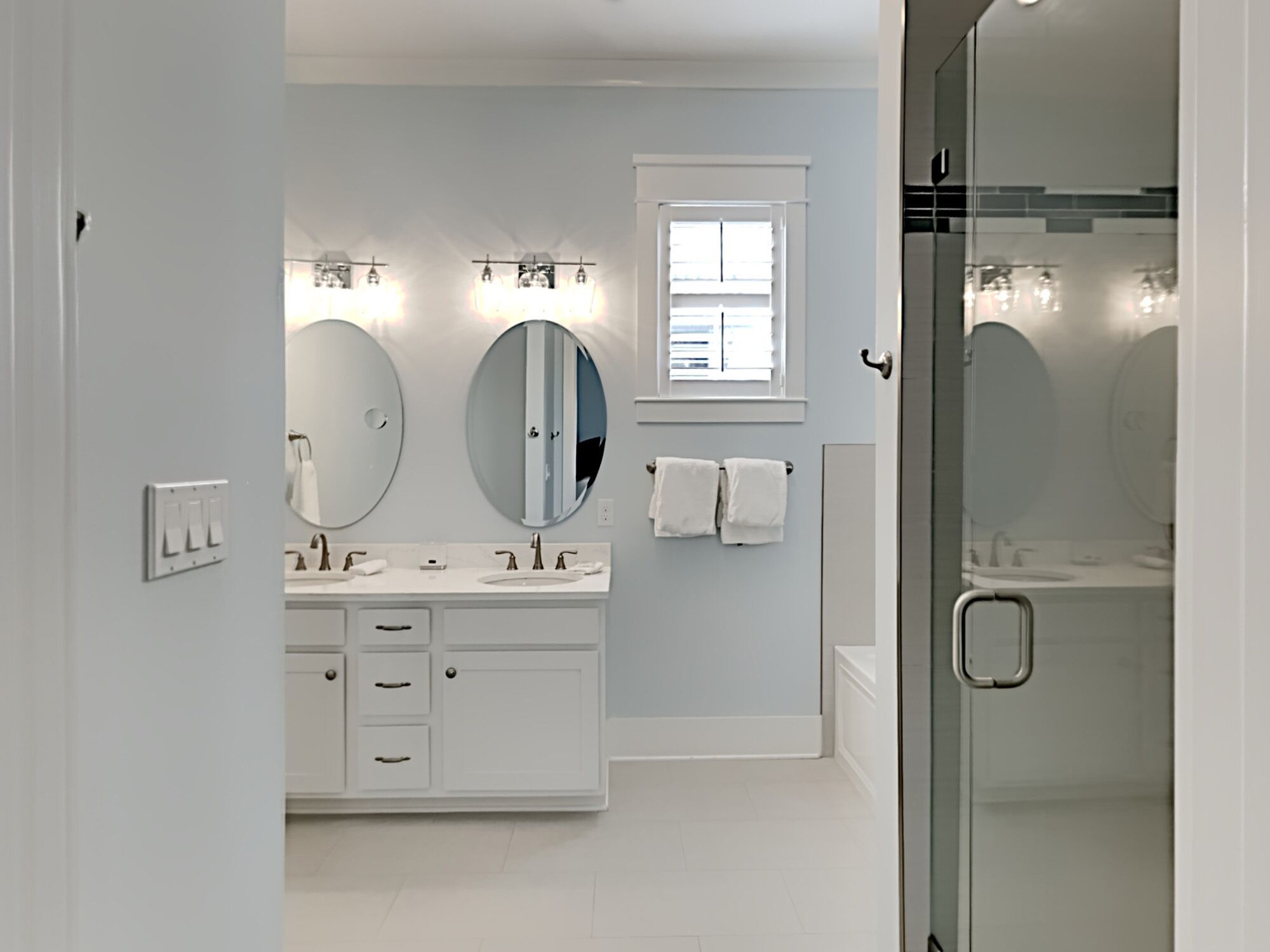 Ein modernes Badezimmer mit einem weißen Waschtisch, zwei großen Spiegeln, einem Fenster und einer Duschkabine aus Glas.
