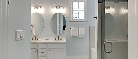 A modern bathroom with a white vanity, two large mirrors, a window, and a glass shower enclosure.