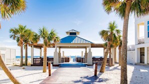 A wooden boardwalk leading to a beach pavilion with a blue roof, surrounded by palm trees and a sandy beach. - Beach Music by Southern Vacation Rentals (Orange Beach)