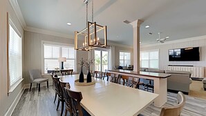 A spacious dining area with a large table and chairs, a modern chandelier, and a kitchen area with a built-in oven and a flat-screen TV. - Beach Music by Southern Vacation Rentals (Orange Beach)