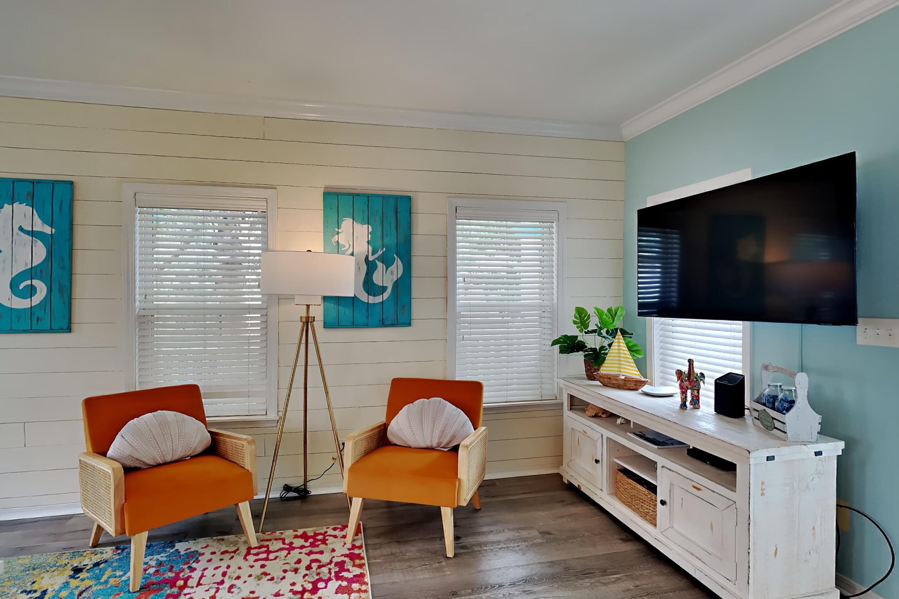 Un salon avec deux fauteuils orange, un meuble de rangement blanc pour appareils électroniques, un téléviseur à écran plat et des œuvres d’art aux tons turquoise accrochées aux murs.