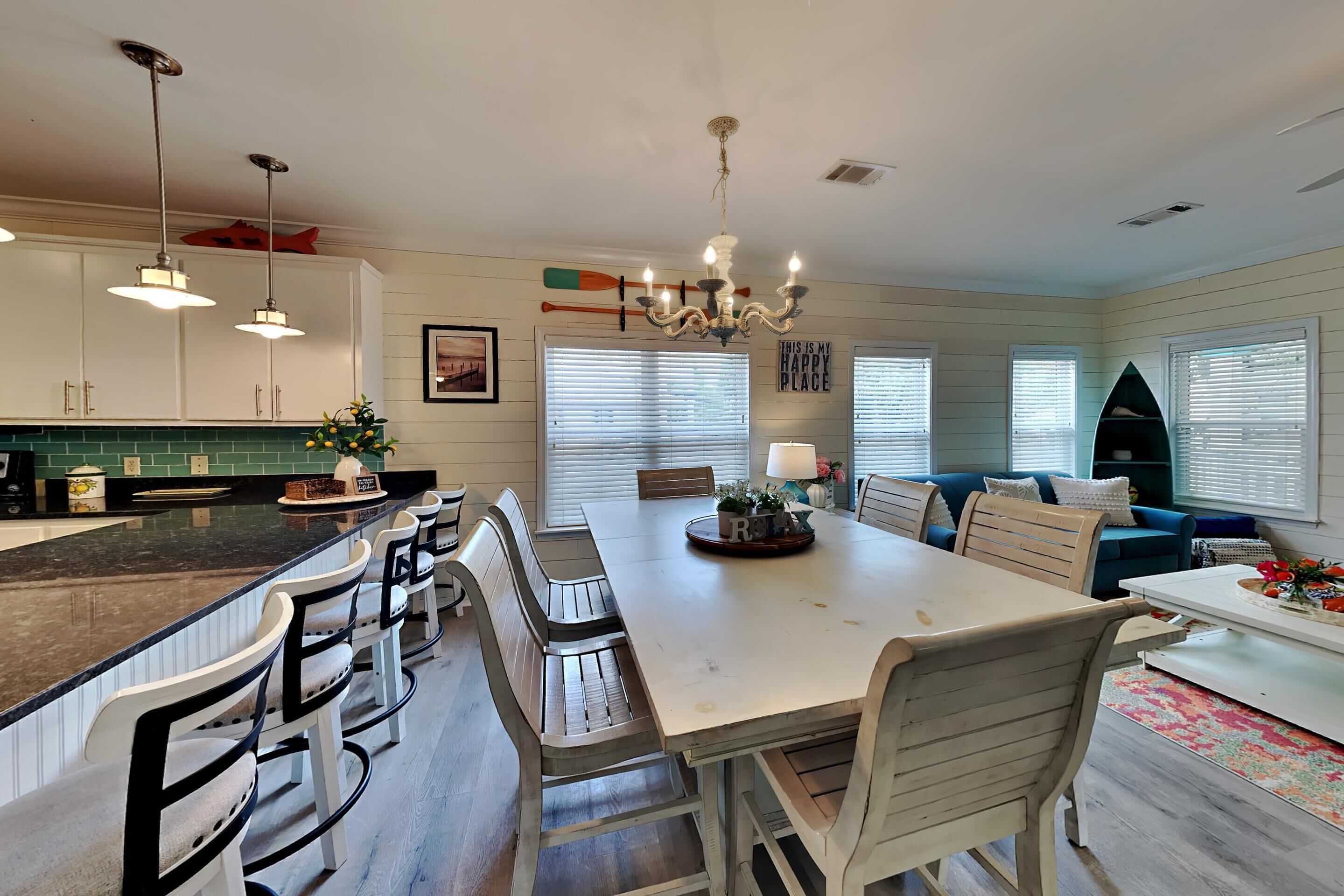 A spacious kitchen with a large island, bar stools, and a dining table. There are pendant lights, a chandelier, and a view of the living room with a couch and a coffee table.