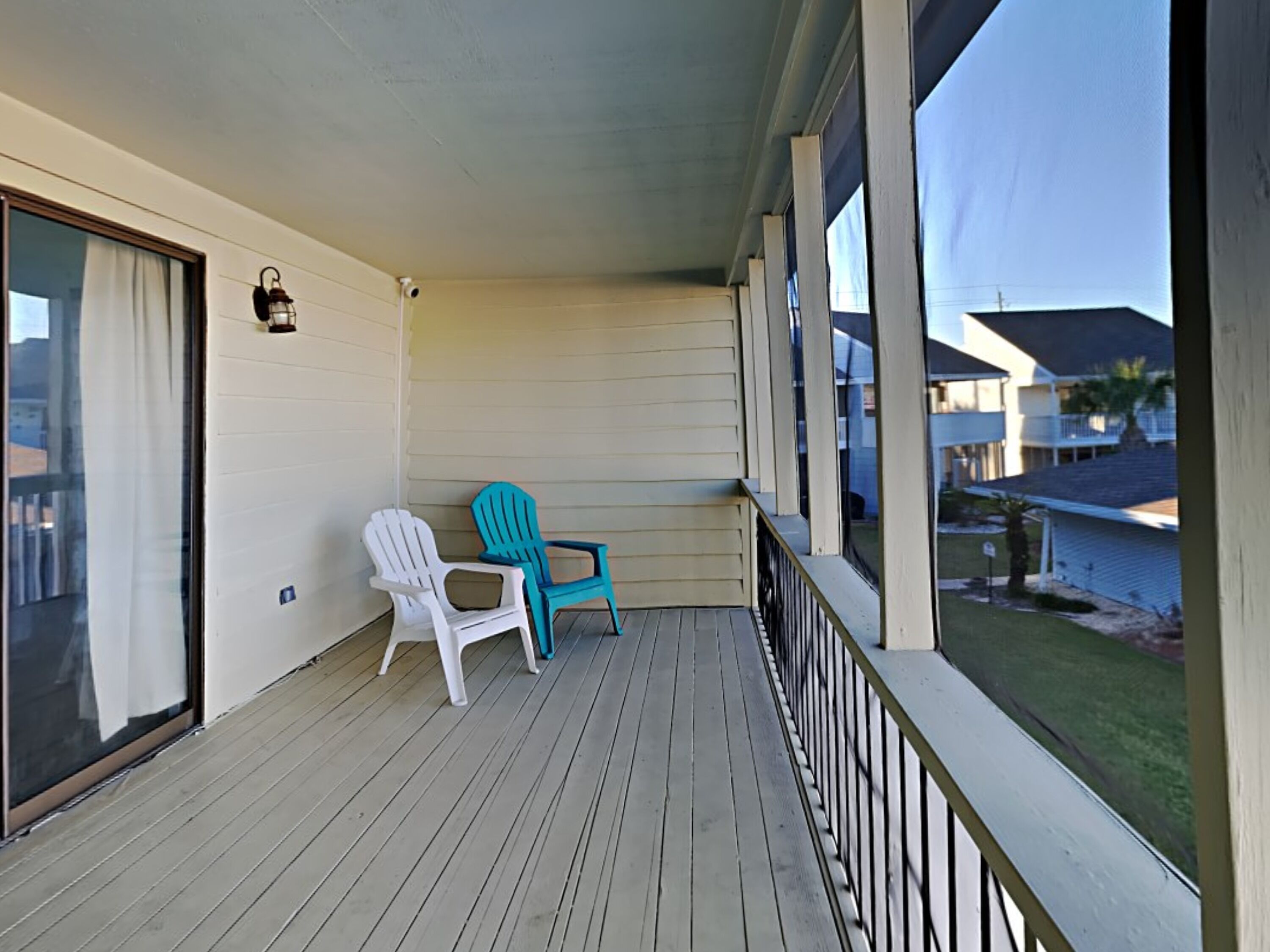 A covered balcony with two Adirondack chairs, a sliding glass door, and a view of a residential area.