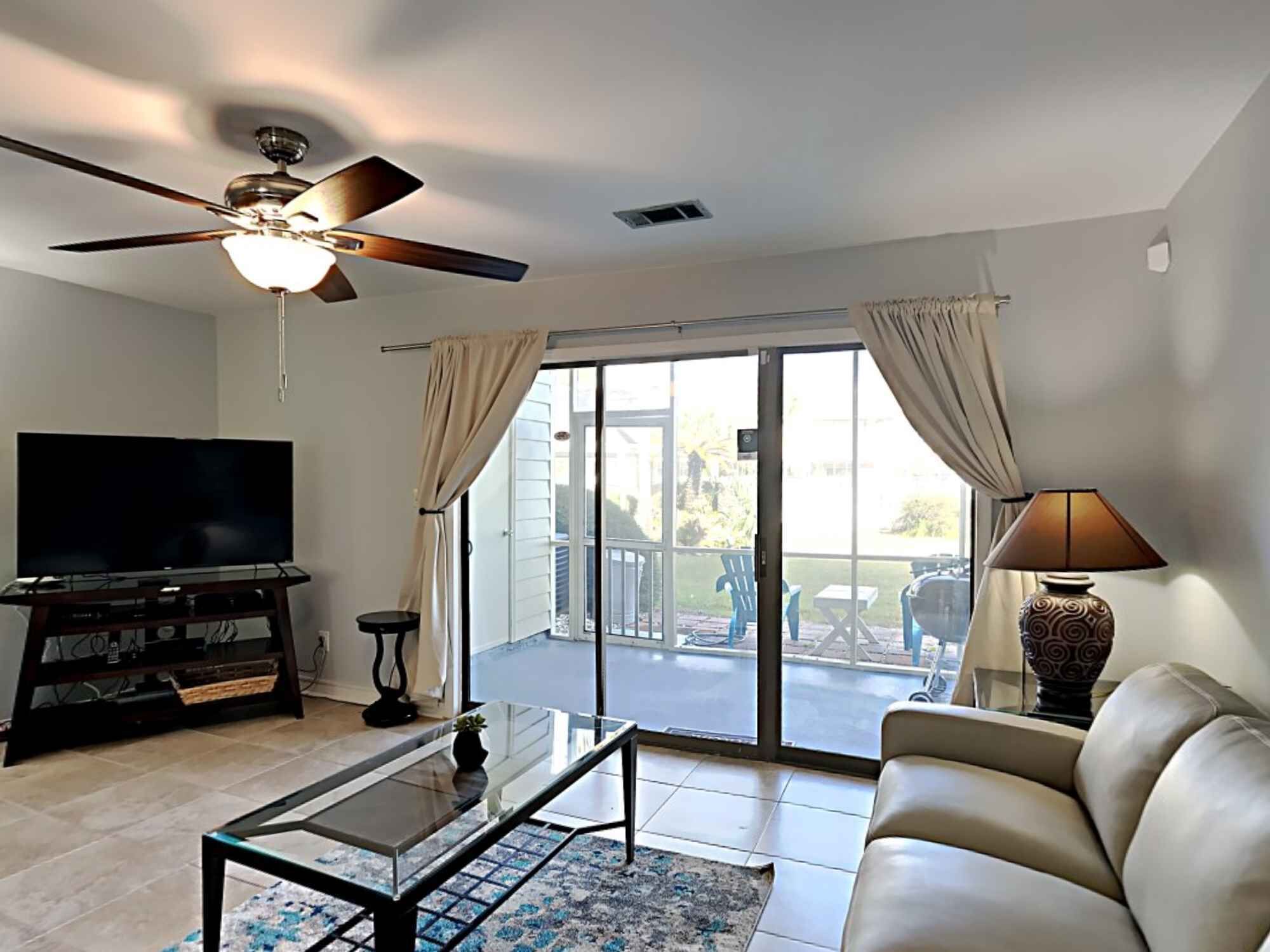 A living room with a flat-screen TV, a glass coffee table, a leather sofa, a ceiling fan, and a sliding glass door leading to an outdoor patio.