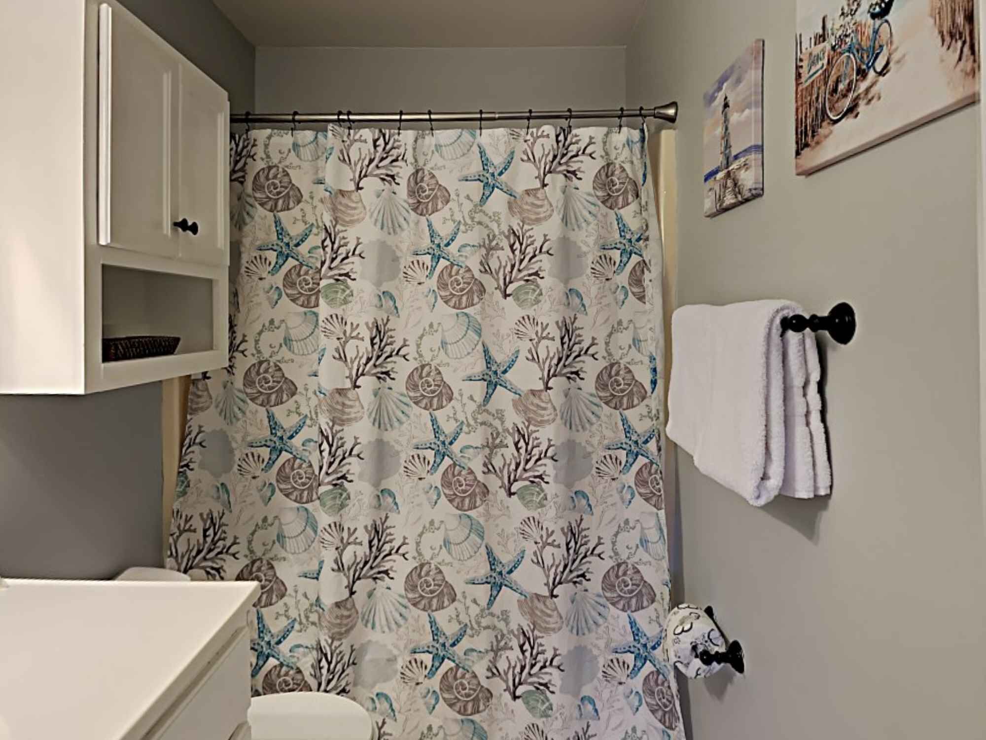 A bathroom with a shower curtain featuring a sea shell and starfish pattern, a white towel hanging on a towel rack, and a small shelf with a basket.