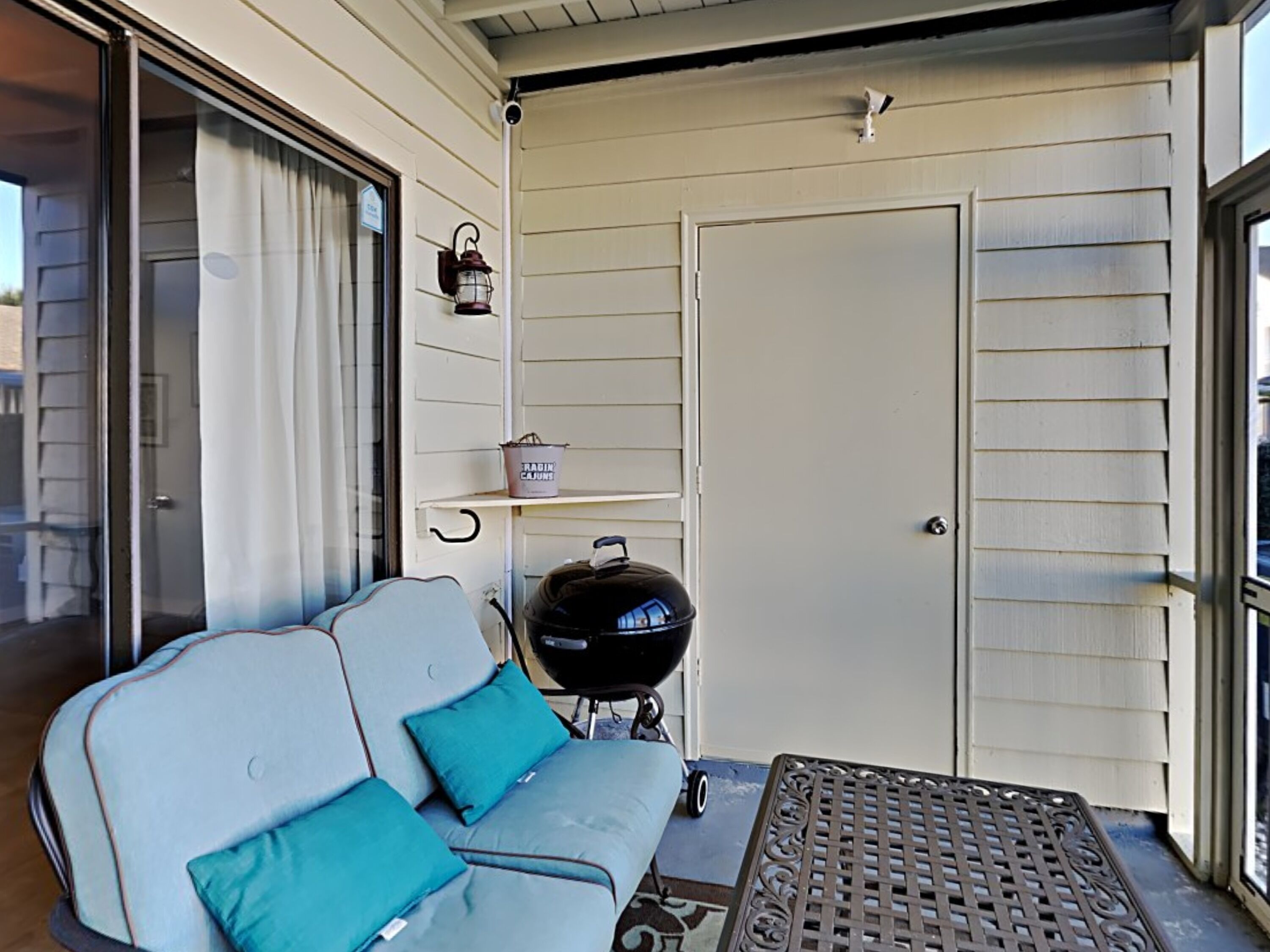A covered porch with a sliding glass door, a white sofa with teal cushions, a barbecue grill, and a metal table.