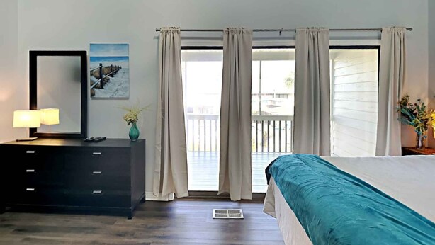 Une chambre à coucher avec un lit, une commode, un miroir, un tableau et une fenêtre avec des rideaux.