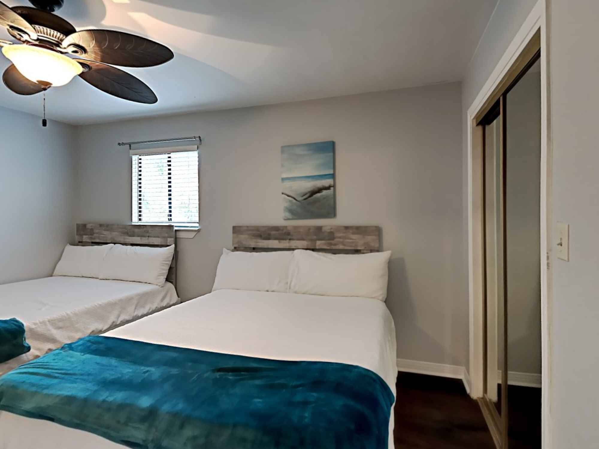 Une chambre avec deux lits, un ventilateur de plafond, une fenêtre avec des stores et un tableau au mur.