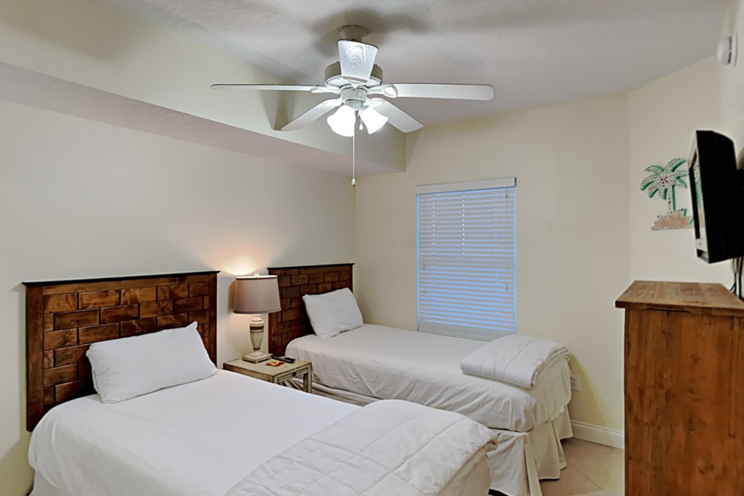 A hotel room with two beds, a wooden headboard, a ceiling fan, a bedside lamp, a window with blinds, and a wooden dresser.