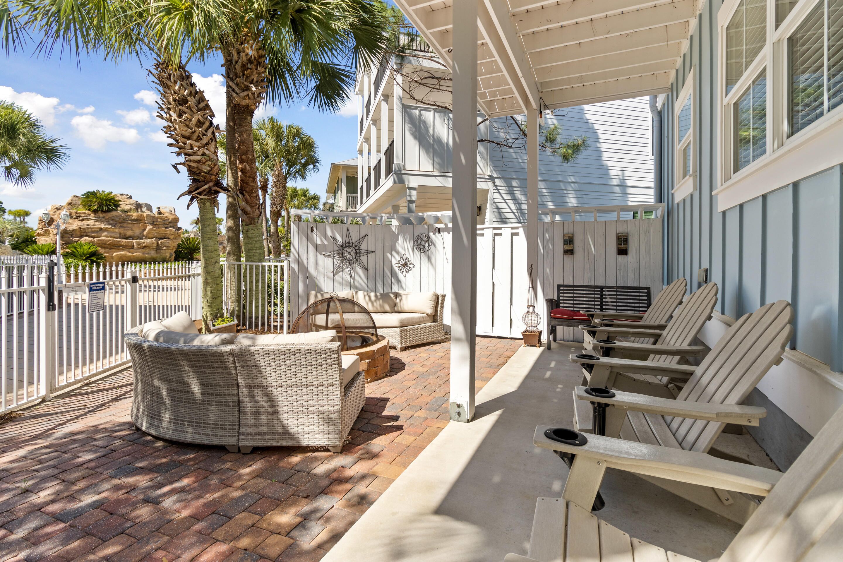 A covered patio with wicker furniture, a fire pit, and a view of a house with a white fence and palm trees.