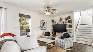 A modern living room with a sofa, armchair, coffee table, and a TV. There's a staircase to the right and a window with blinds. - Easy Breezy by Southern Vacation Rentals (Santa Rosa Beach)