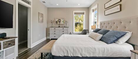 A bedroom with a large bed, a ceiling fan, a dresser, a TV, and a window with a view of greenery.