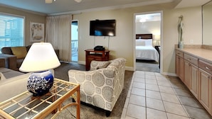 A hotel room with a ceiling fan, a TV, a bed, a kitchenette, and a seating area with a lamp and a patterned chair. - Spinnaker #2A by Southern Vacation Rentals (Pensacola)