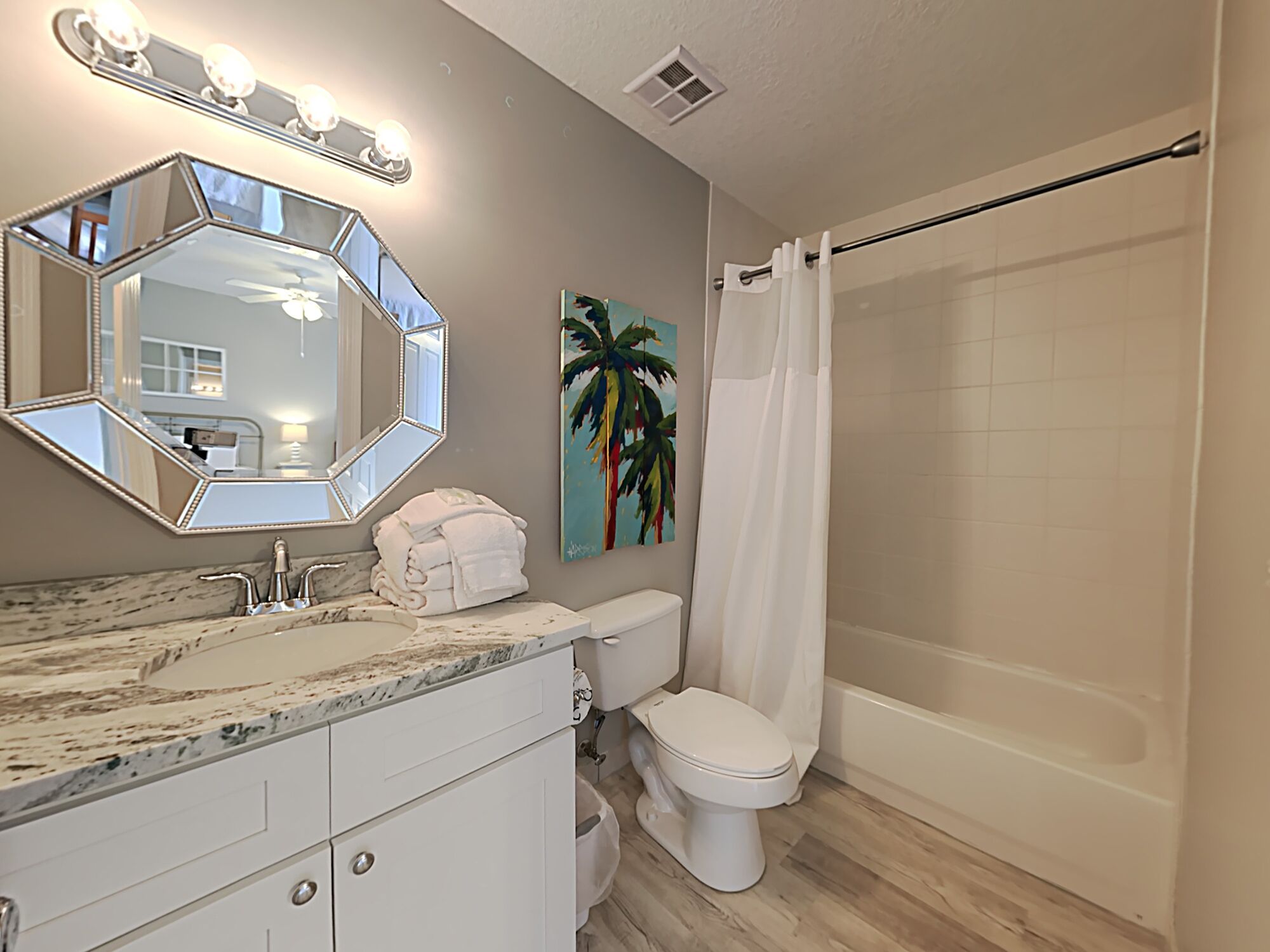 A bathroom with a marble vanity, a toilet, a shower with a curtain, and a large mirror with lights.