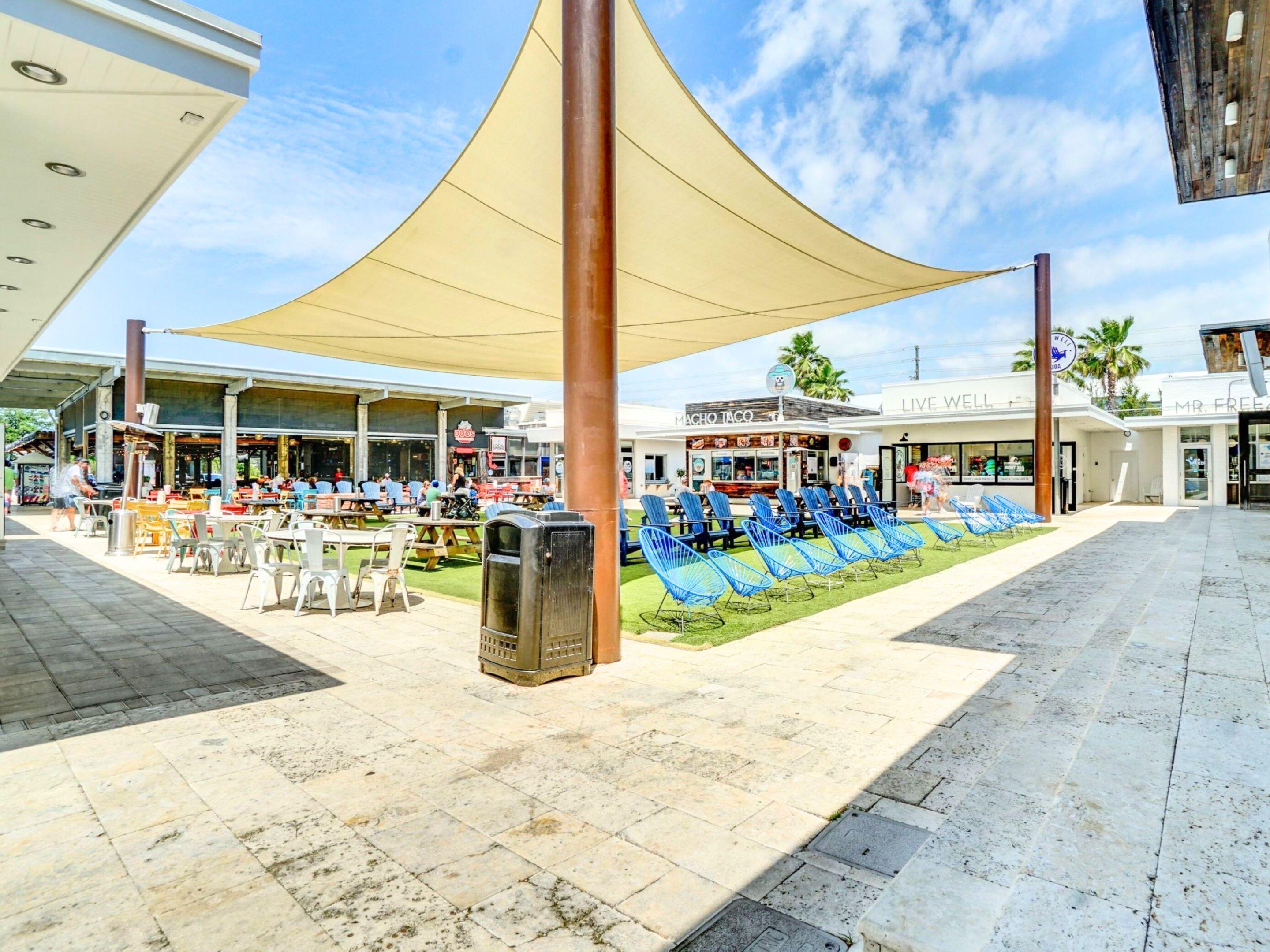 An outdoor seating area with tables and chairs, a trash bin, and a shaded area with a yellow canopy.