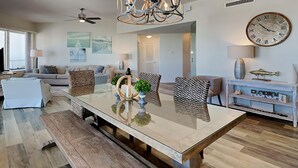 A spacious living room with a large glass-top dining table, a chandelier, and a wall clock.