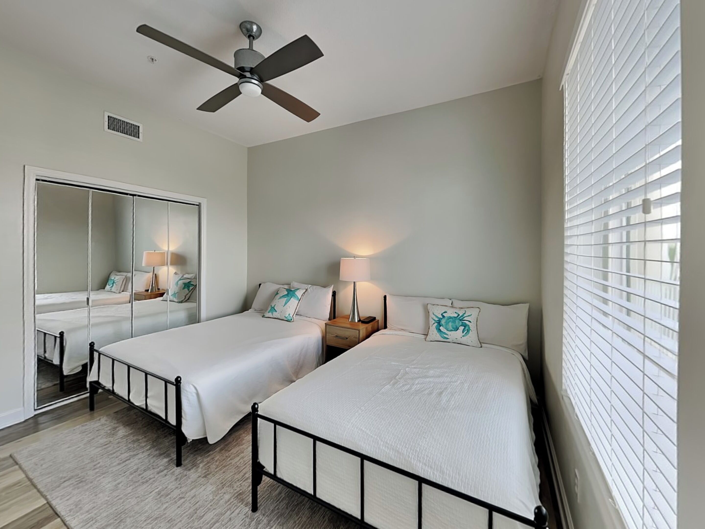 A hotel room with two single beds, a ceiling fan, a mirror, and a window with blinds.