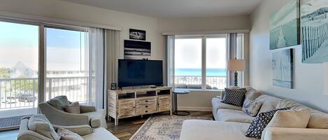 A spacious living room with a large TV, comfortable seating, and a balcony with a view.