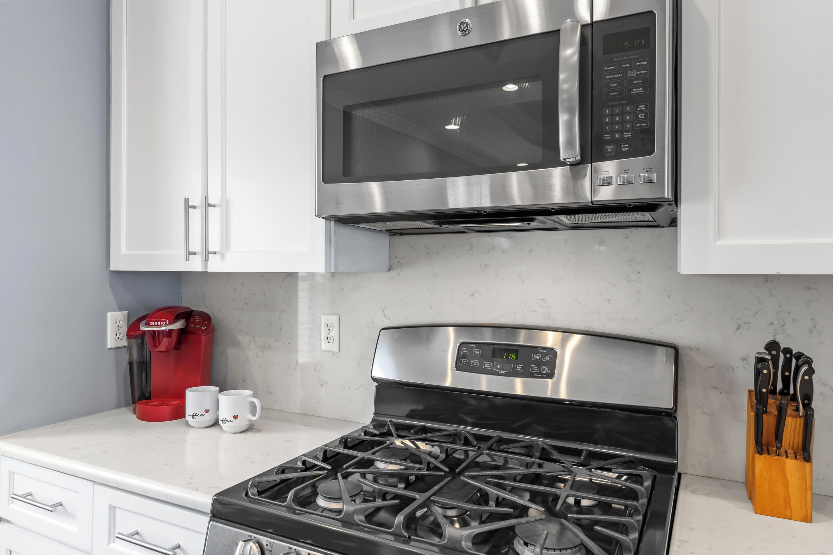 A kitchen with white cabinets, a stainless steel microwave, a red coffee maker, and a gas stove.