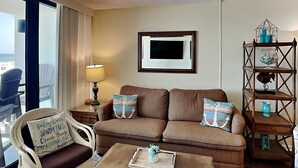 Een woonkamer met een bruine bank, een witte fauteuil, een bijzettafeltje, een lamp, een lijst met een afbeelding en een houten plank met decoratieve spullen.