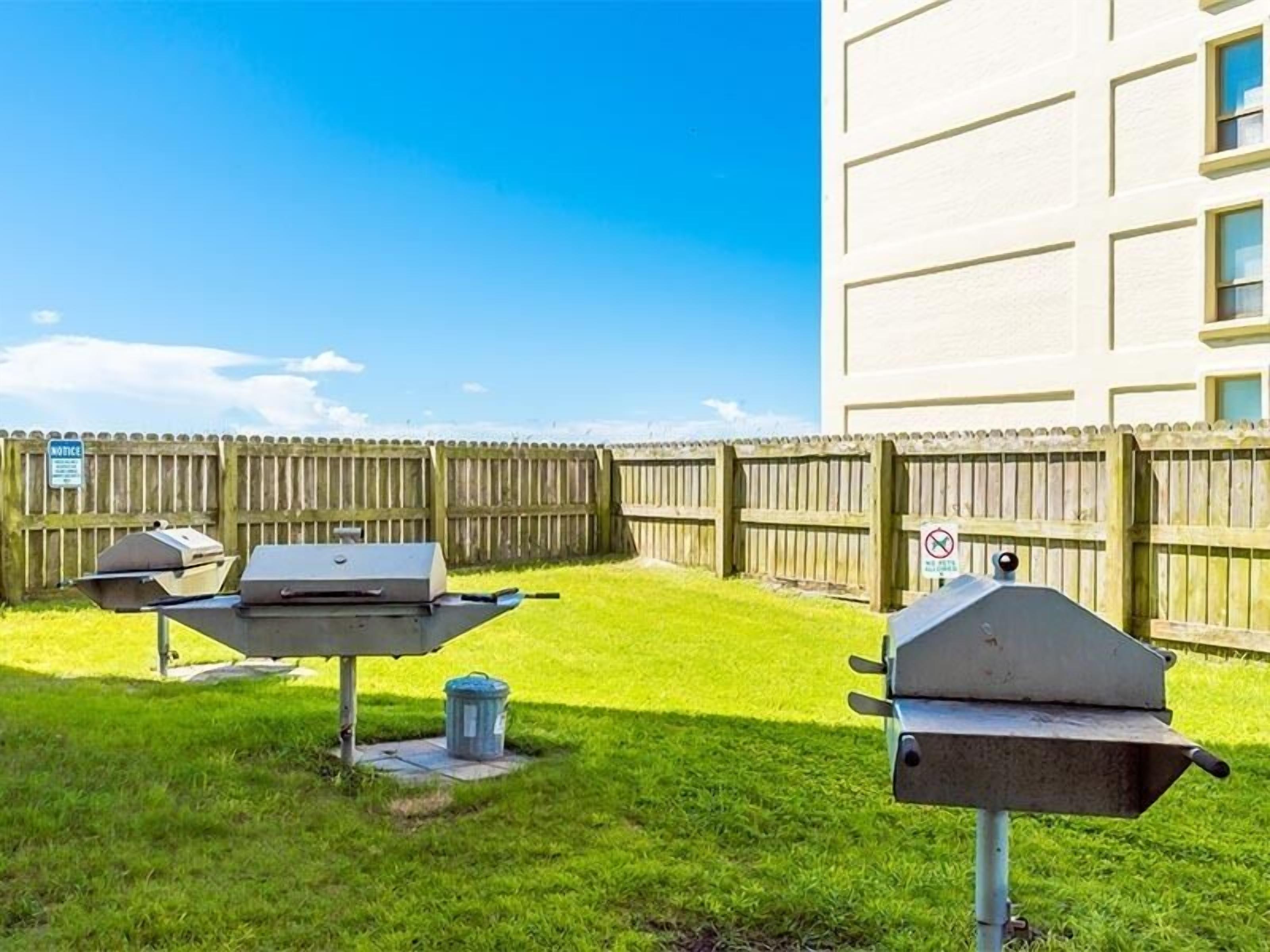 A backyard with two barbecue grills, a wooden fence, and a trash can.