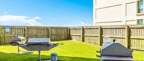 A backyard with two barbecue grills, a wooden fence, and a trash can.