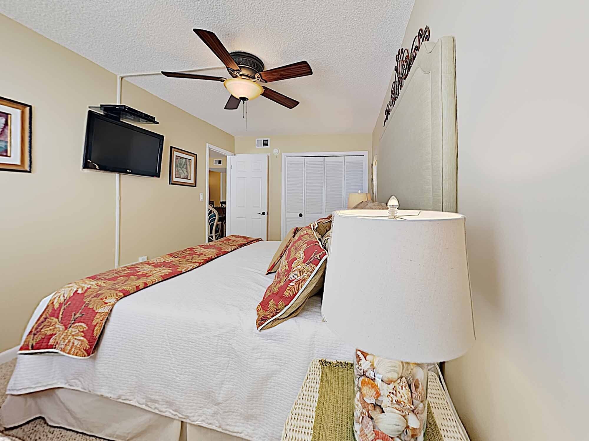 A hotel room with a bed, a ceiling fan, a TV, and a lamp.