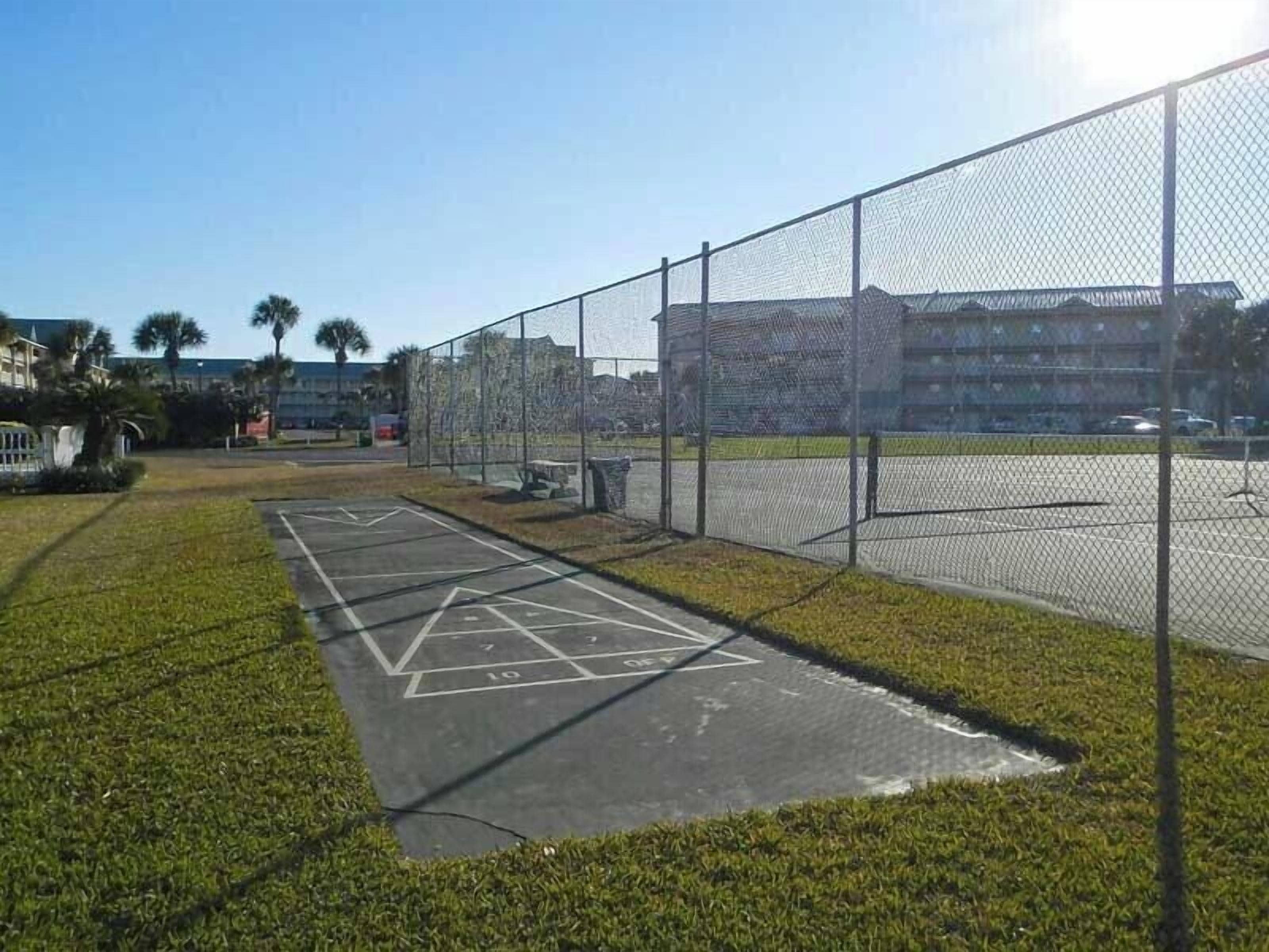 A tennis court with a net, surrounded by a chain-link fence, and a parking lot in the background.