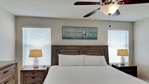 A bedroom with a large bed, two bedside tables with lamps, a ceiling fan, and a painting of sailboats on the wall. - The Beach Is Our Happy Place by Southern Vacation Rentals (Miramar Beach)