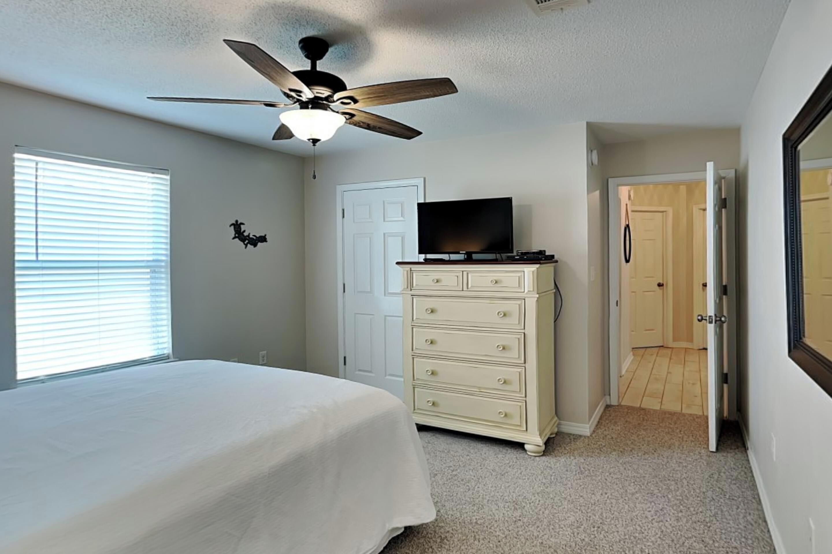 Een slaapkamer met een bed, een dressoir, een plafondventilator, een televisie en een deur naar een andere kamer.